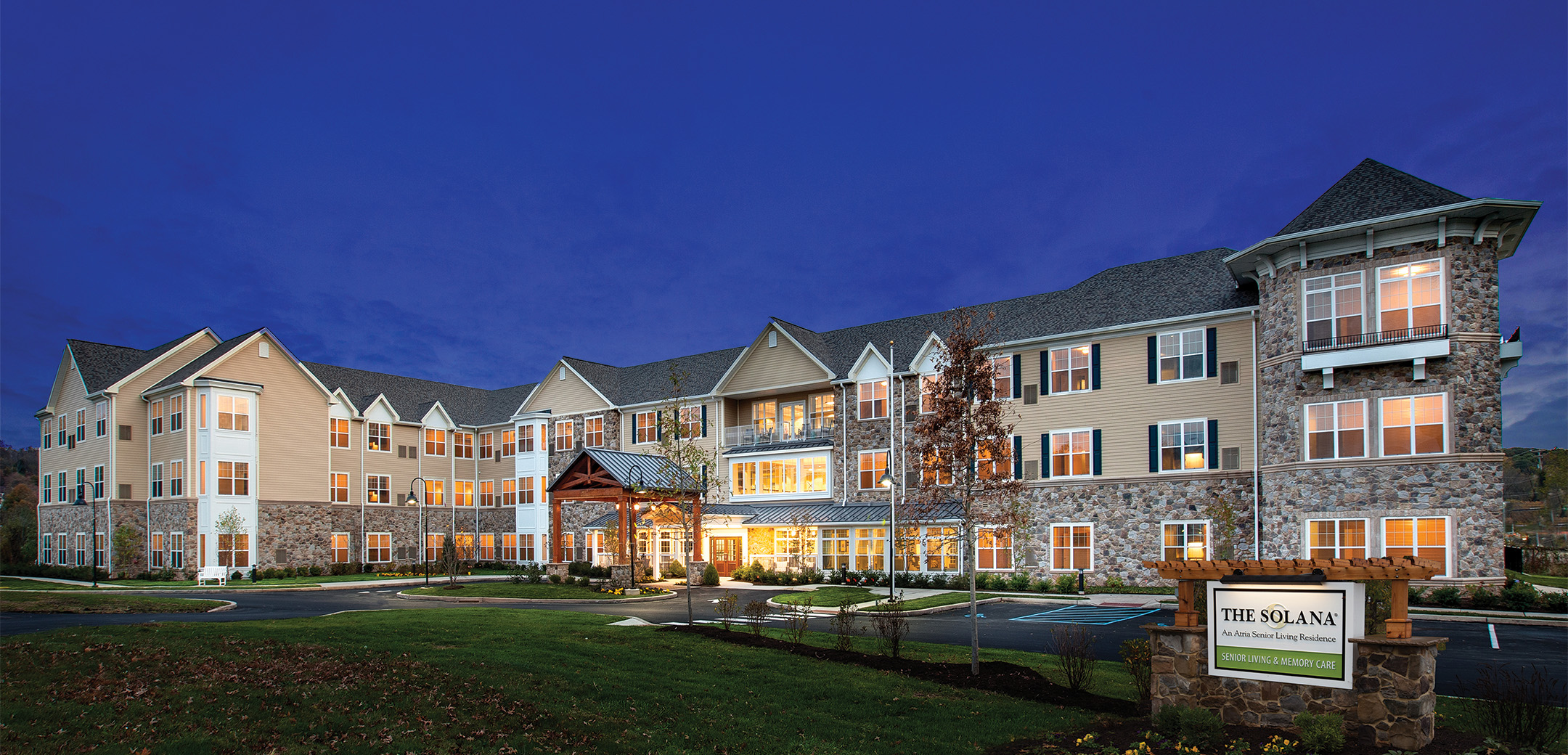 An exterior nighttime view of the Formation- Shelbourne stone brick and grey shingle roofing with a driveway, parking lot and grass lawn in the foreground.
