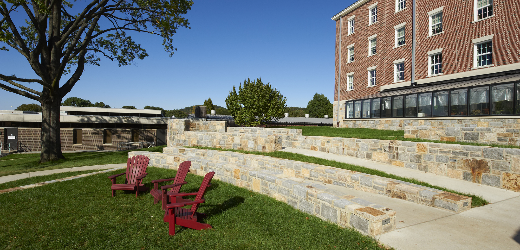 A close up view of the Merner Pfeiffer Hall of Science inner courtyard, showcasing the accessibility ramp, background tree and grass lawn with red chairs.
