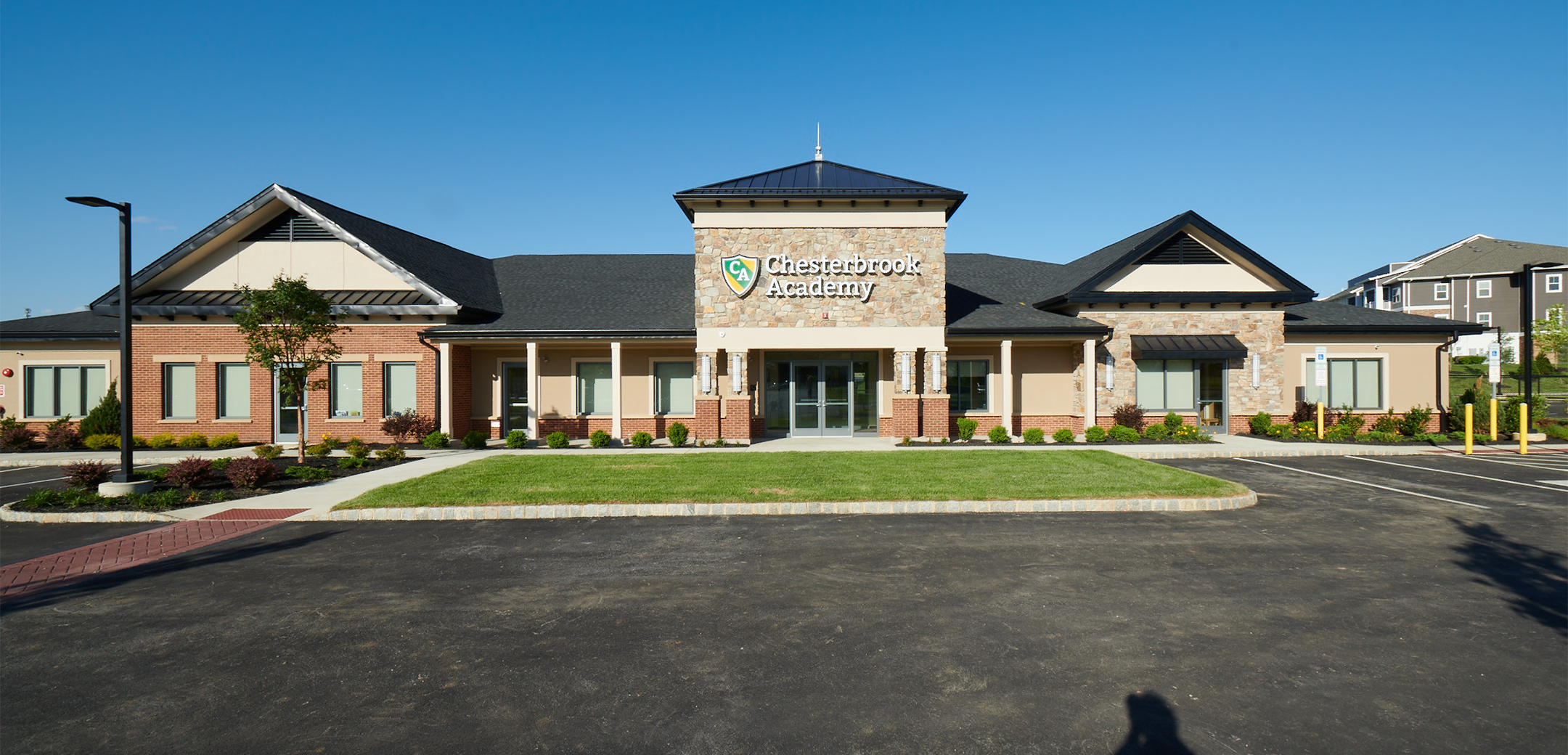 A front view of the single story Chesterbrook Academy building showcasing the front green lawn area and parking lot in front of the entrance
