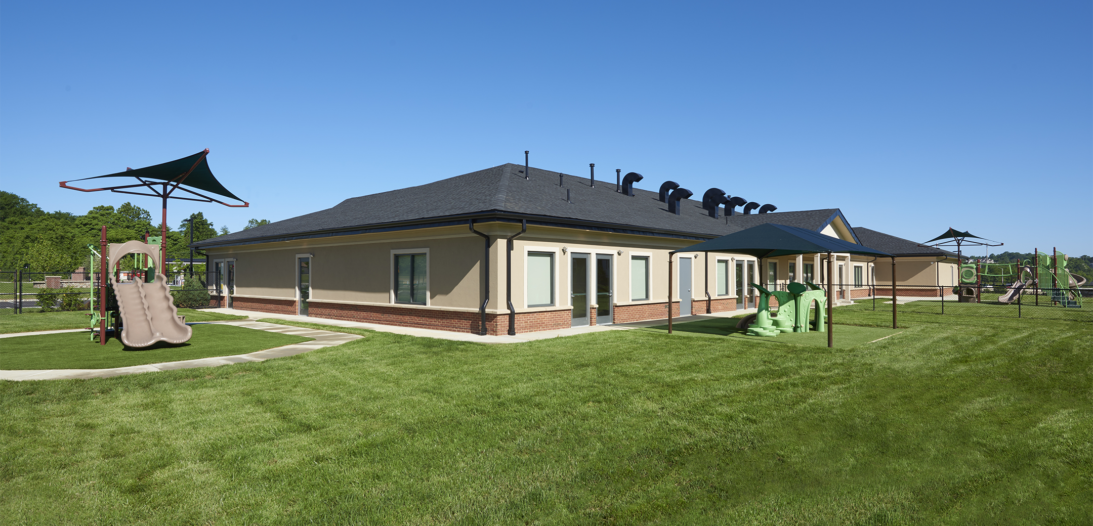 An angled view of the back side of the single story Chesterbrook Academy building showcasing the large green grass yard with a children's playground.
