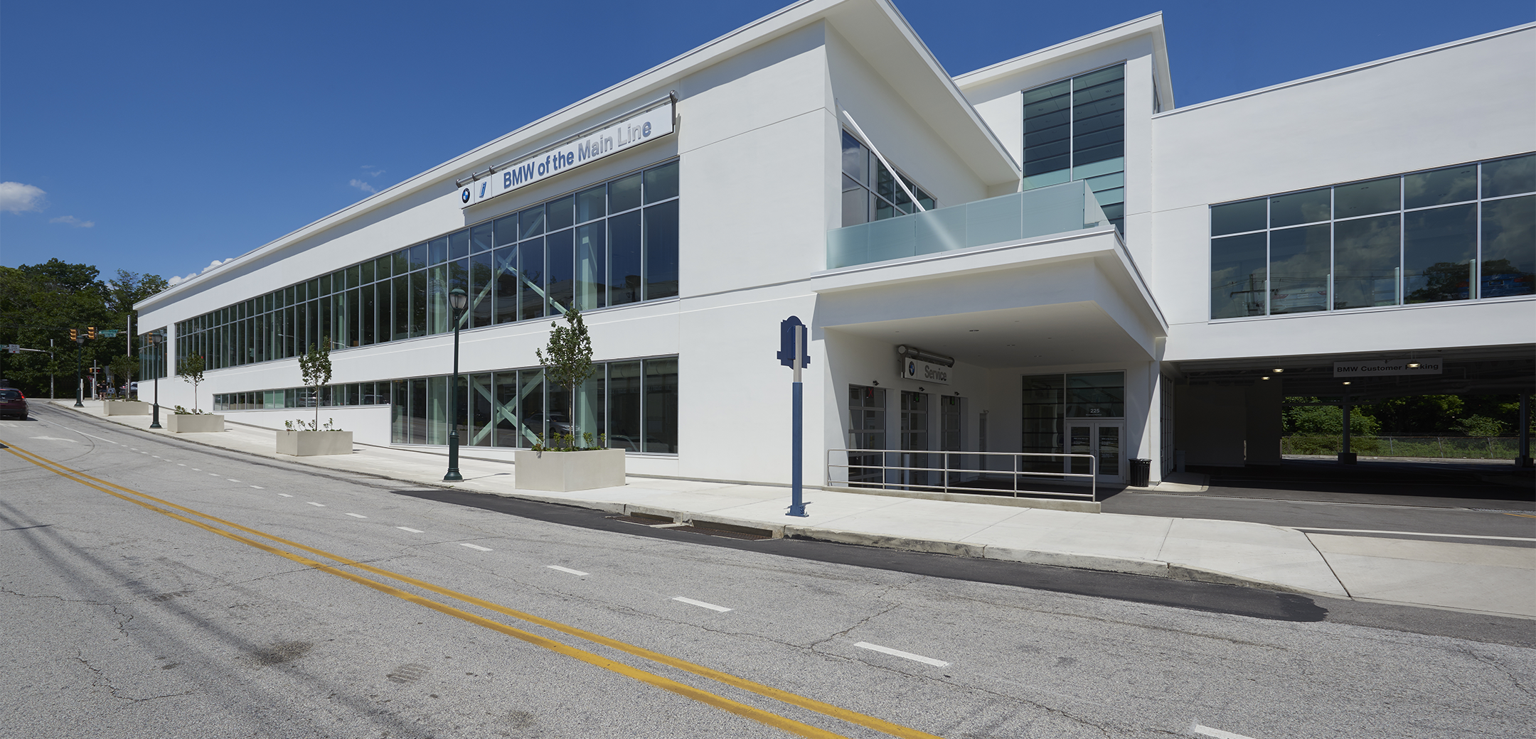 An exterior view of the Main Line BMW two story grey concrete building with glass windows along the full length of the walls and a road in the foreground.