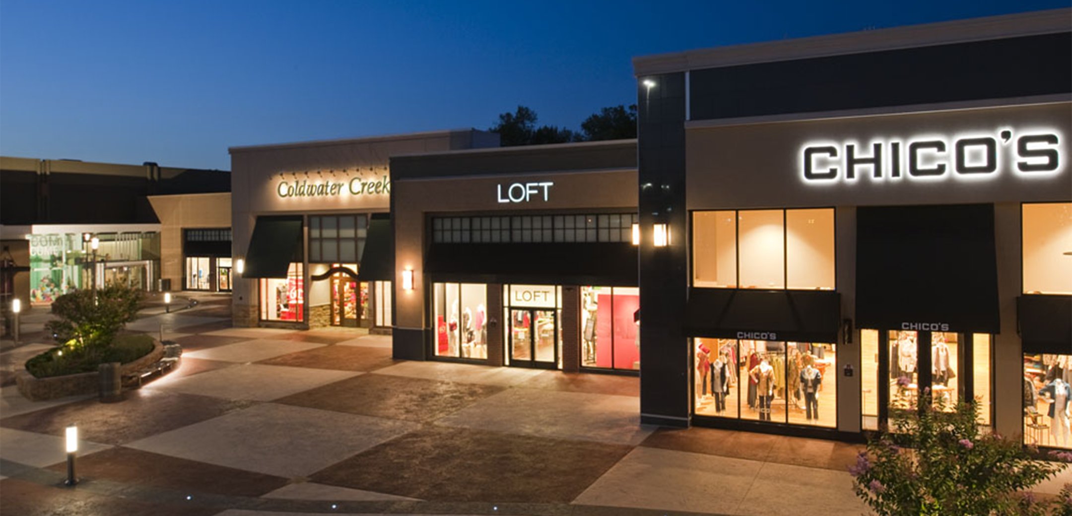 An exterior nighttime view of the Plymouth Meeting Mall showcasing the courtyard, shopfronts and plant islands with seating areas in the middle of the walkways.