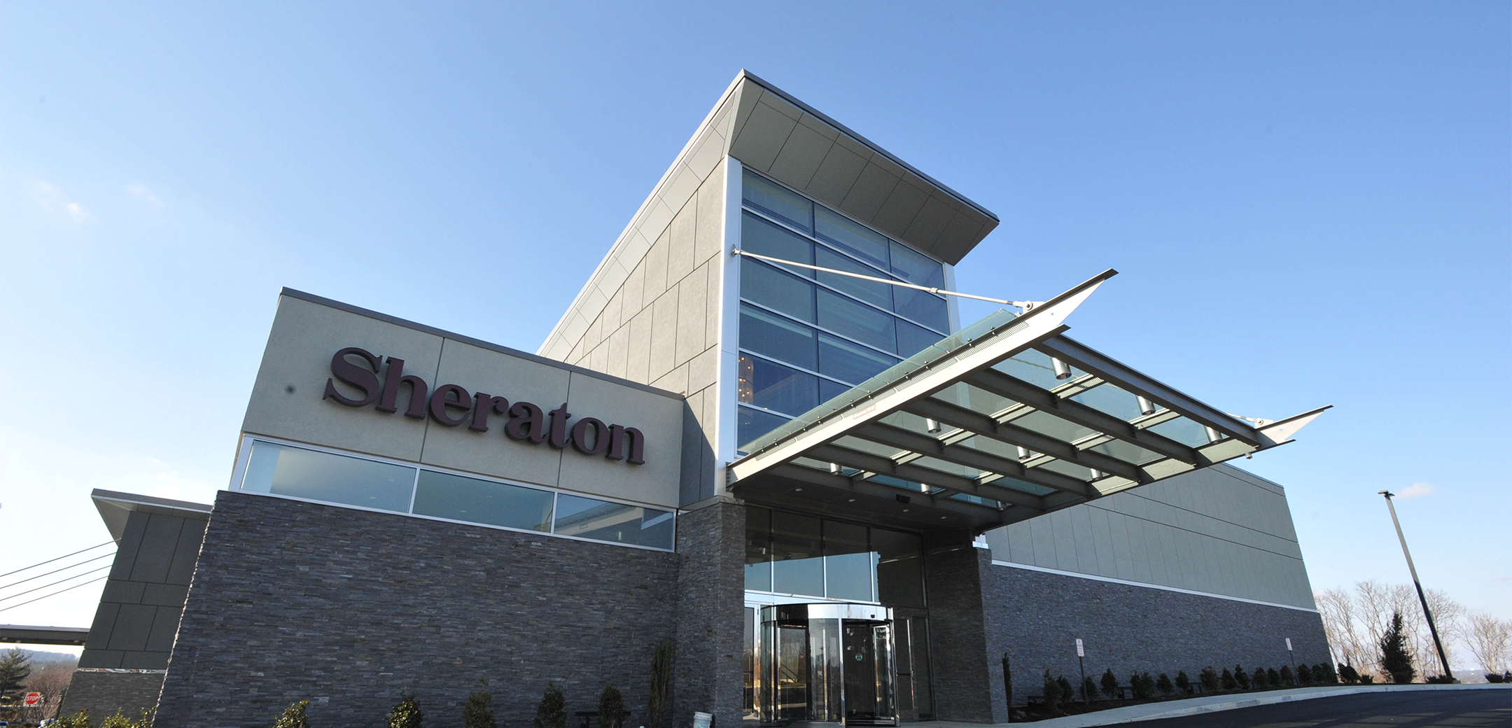 A close up of the Sheraton Valley Forge driveway entrance showcasing their logo and glass overhang above rotating doors.