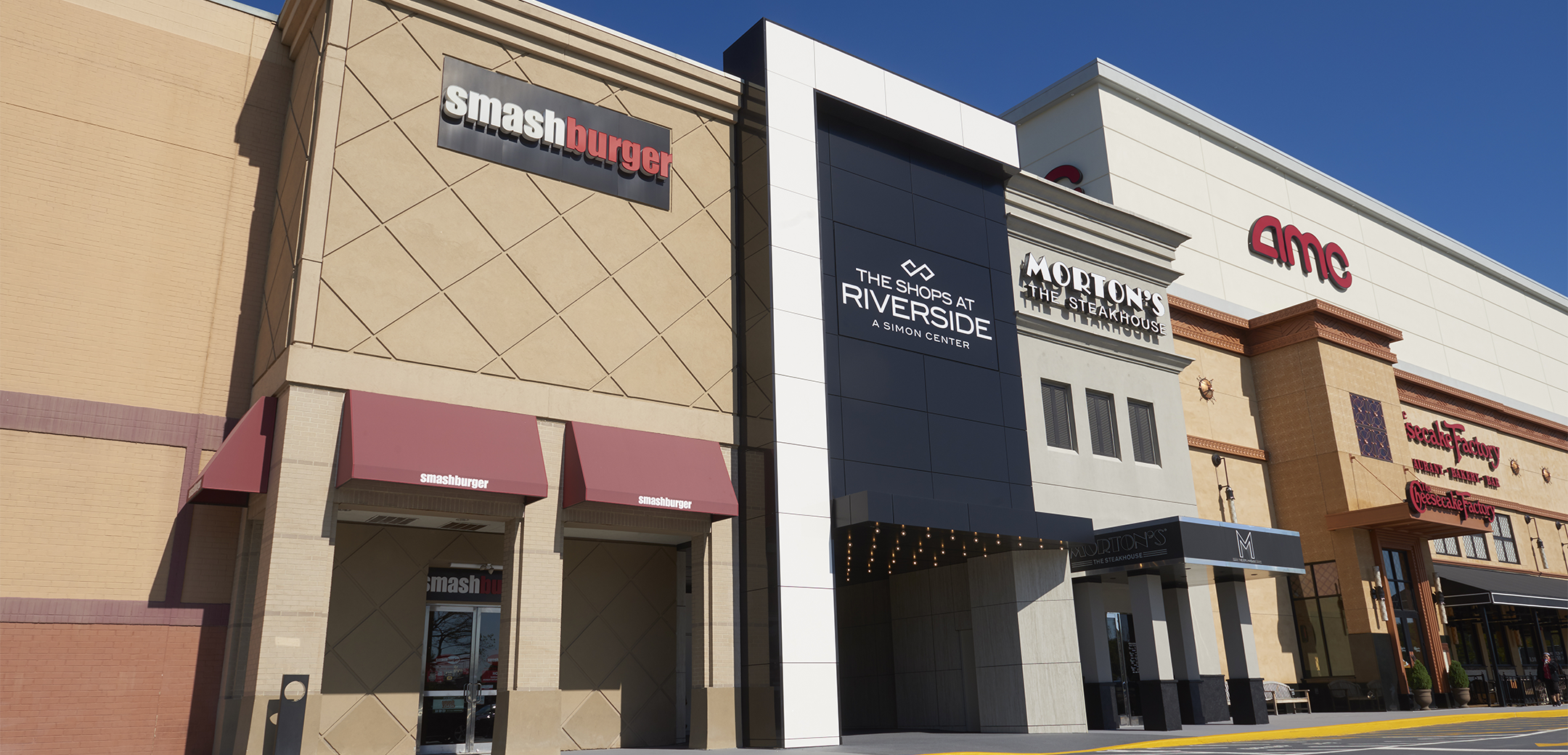 An exterior view of the Shops at Riverside building strip, showcasing the shop signages, navy blue accented overhang panel entrance and streetside parking.