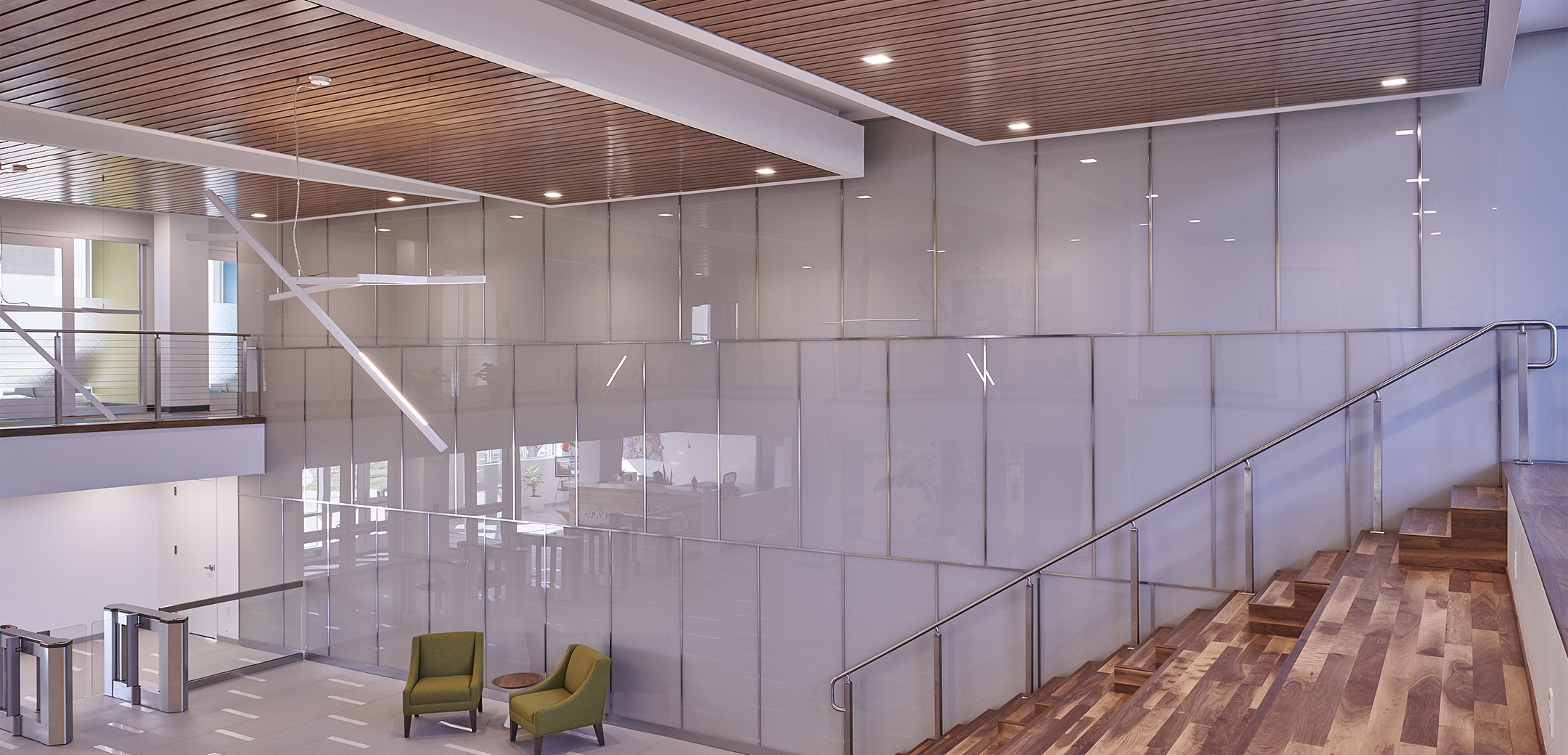 An angled view from the wooden grandstand style seats overlooking the first floor lobby with office furniture, office sercurity gates, and the side view of the staircase against glass walls.