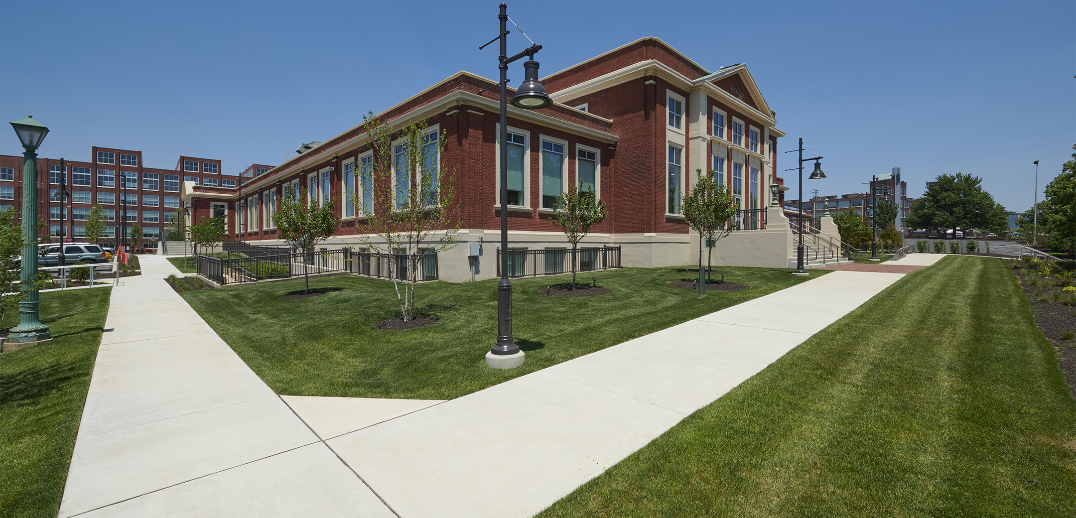 An angled view of a brick building with white sidewalks and landscaping in the front and the Knitting Mills mixed-campus buildings in the background.