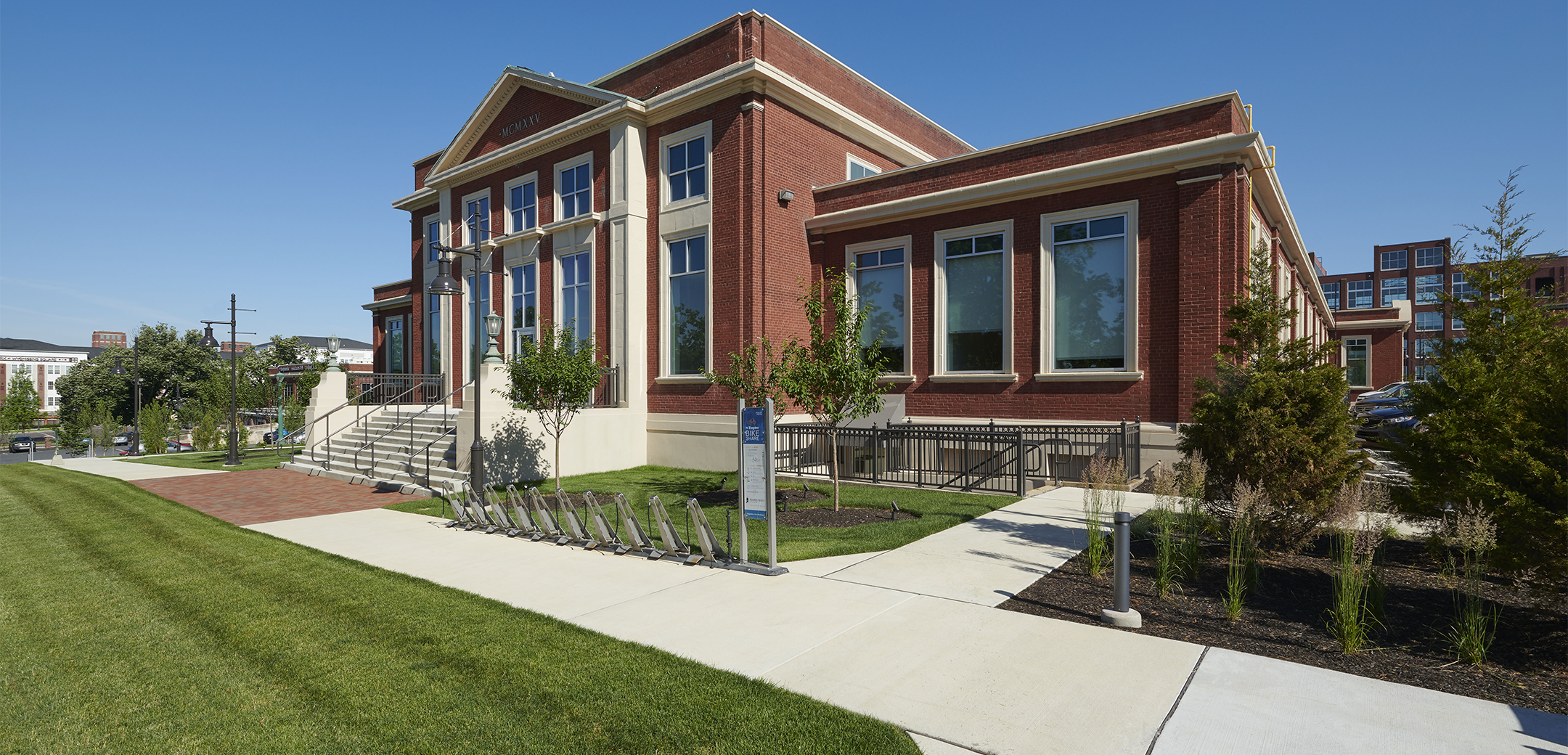 An angled view of a brick building that features bike racks, white sidewalks and landscaping in the front and the Knitting Mills mixed-campus buildings in the background.